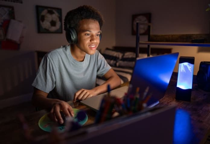 A focused high school student wearing headphones is sitting at a desk and looking at his laptop screen, representing a student engaged in online virtual school learning.