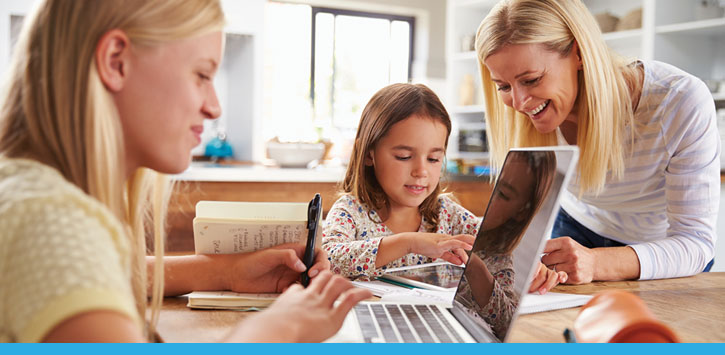 Two younger students at a dining room table, learning on their laptops, with the support of their mom.