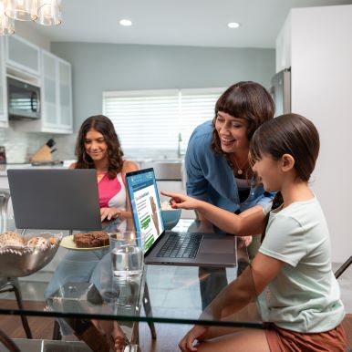 Family around the kitchen table with mother assisting younger daughter with her work on the daughter's laptop