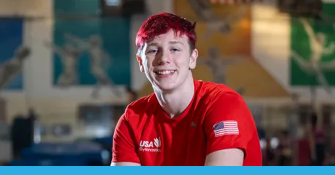 FLVS Student Shines at World Trampoline Gymnastics Championships West Fowler wearing a red USA Gymnastics shirt with an American flag patch, seated in an indoor gymnastics facility with colorful wall murals in the background.