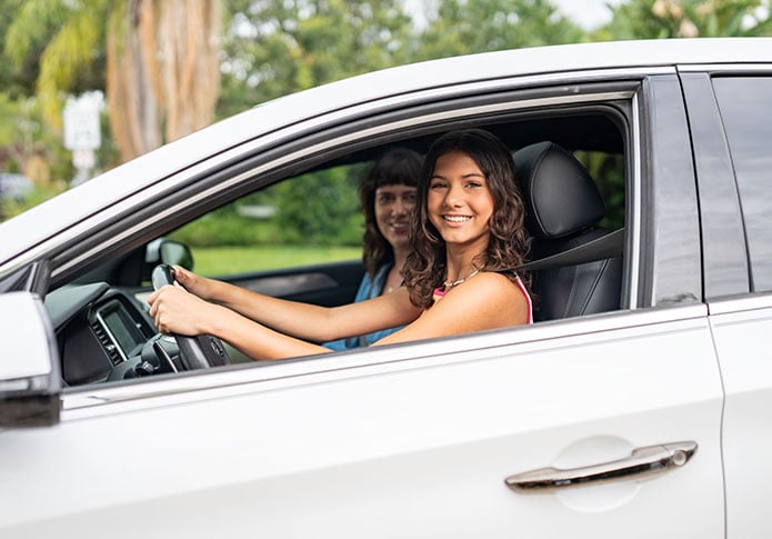 Teenage girl smiling while driving a car, with an adult instructor or parent in the passenger seat, illustrating the practical application of skills learned through FLVS's online Driver Education course.