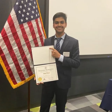 Sarthak Gupta stands in front of American flag holding his certificate announcing his win