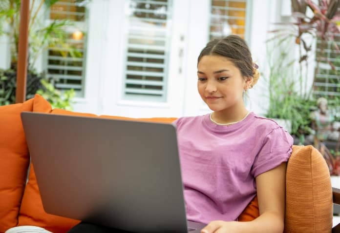 Teenage girl comfortably studying on a laptop outdoors on a patio sofa. This image highlights the flexibility and anytime, anywhere access of the FLVS Flex individual courses.
